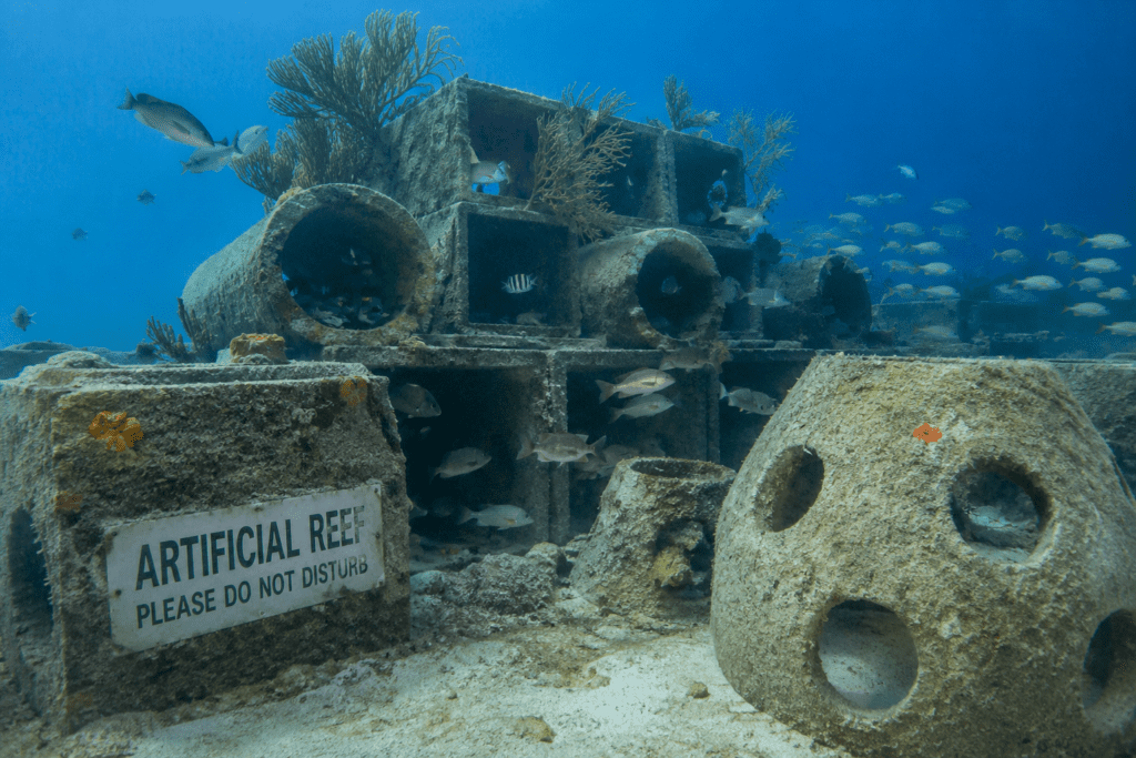 Artificial reef structure underwater featuring concrete reef modules, pipes, and marine growth surrounded by schools of fish in clear blue Gulf waters.