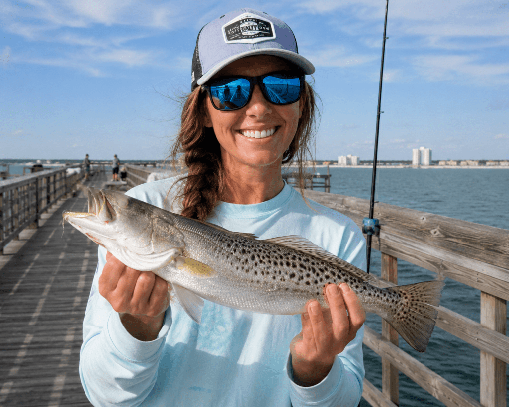 Smiling fisherwoman standing on a Gulf Coast fishing pier holding a freshly caught speckled trout. The calm coastal waters, fishing rod, and distant shoreline create a classic Mississippi Gulf Coast fishing scene.